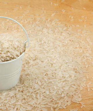 A pile of spilled rice on a wooden surface, a small white tin buck of rice in the centre