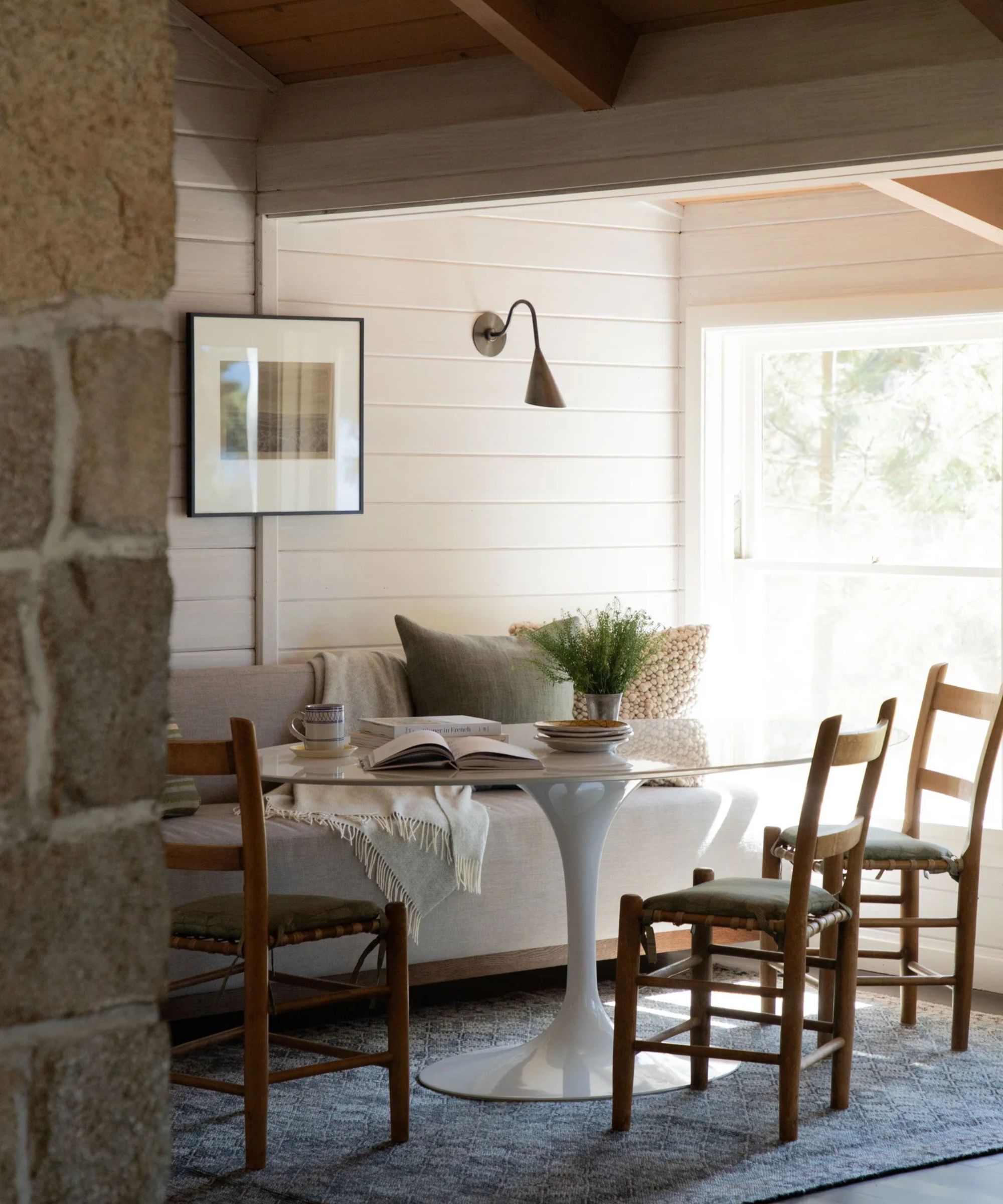 A cozy dining nook with paneled walls, banquette seating, and white oval pedestal table, wooden chairs, and an antique painting in a modern frame displayed on the wall