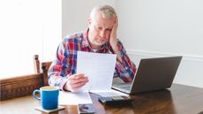 An older man looks stressed as he looks at paperwork while sitting at his dining room table.