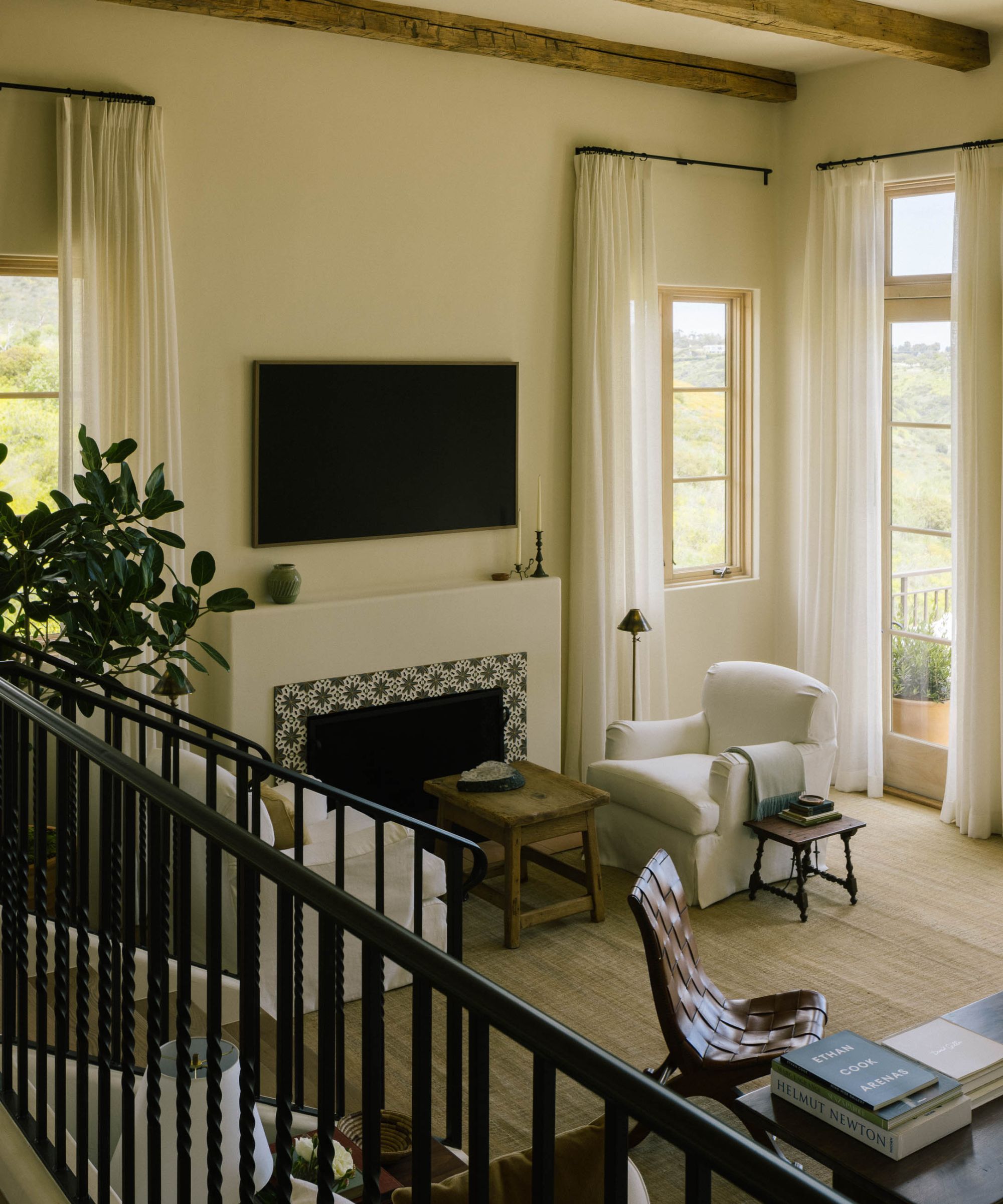 a neutral living room with soaring ceilings, rustic beams, wrought iron staircase, a Spanish-style fireplace with painted tiled and casual linen covered chairs