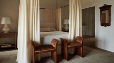 a calm neutral bedroom with limewashed walls, a black metal four poster bed with cream curtains, a neutral rug, and two chocolate brown ottomans at the foot of the bed with antique wood mirror and nightstand
