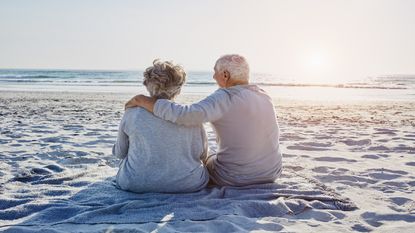 A senior retired couple sit on a beach.