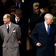 Prince Edward wearing a gray tweed coat looking to one side standing next to Prince Andrew wearing a navy coat looking the other direction