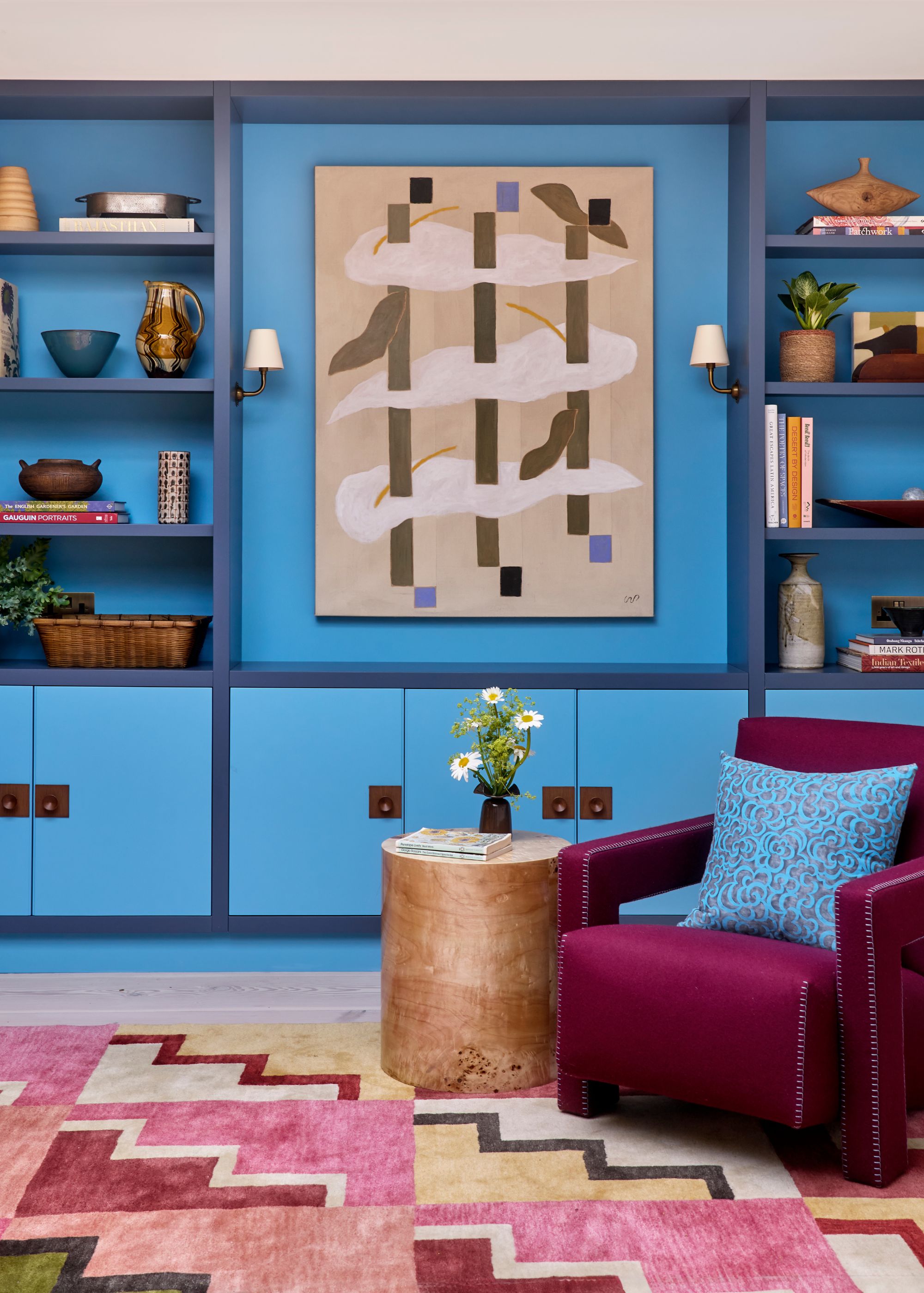 A cozy living room with bright blue color-drenched walls with cabinets and shelves, with dark blue edges. A burgundy armchair and side table, and a pink, red, and beige geometric-print rug.