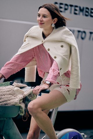 A NYFW SS25 Street Style guest wearing white shorts, a red and white striped button-down shirt, white lady jacket on a bike