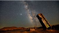 A bus is photographed balanced on its end in the desert as the Milky Way arches overhead at night