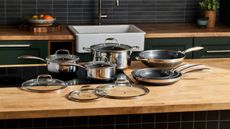 A complete set of stainless steel Hexclad hyrid pans on a wooden kitchen counter with a black stove top set into it. 