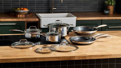 A complete set of stainless steel Hexclad hyrid pans on a wooden kitchen counter with a black stove top set into it.