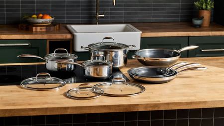 A complete set of stainless steel Hexclad hyrid pans on a wooden kitchen counter with a black stove top set into it. 