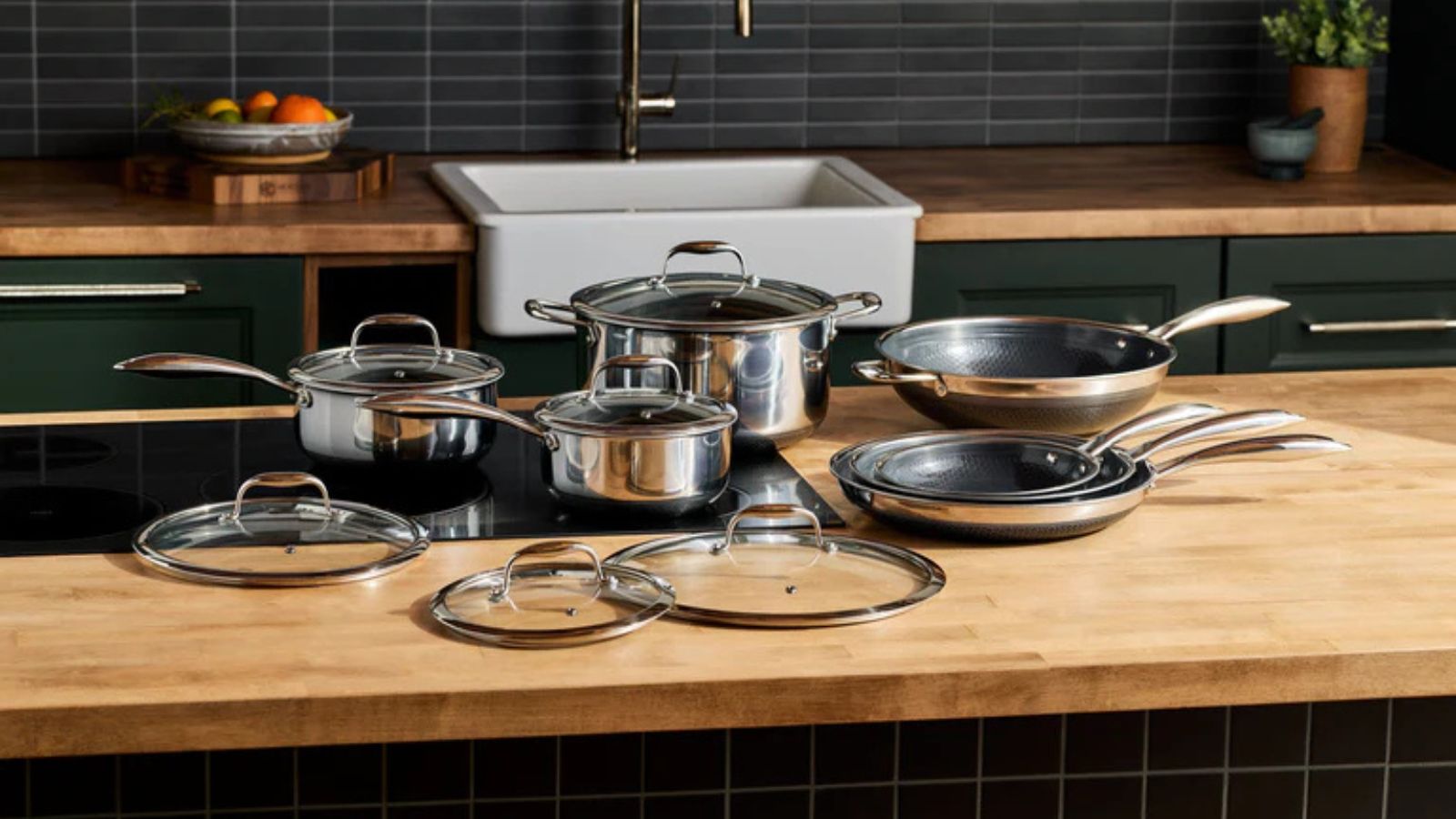 A complete set of stainless steel Hexclad hyrid pans on a wooden kitchen counter with a black stove top set into it. 