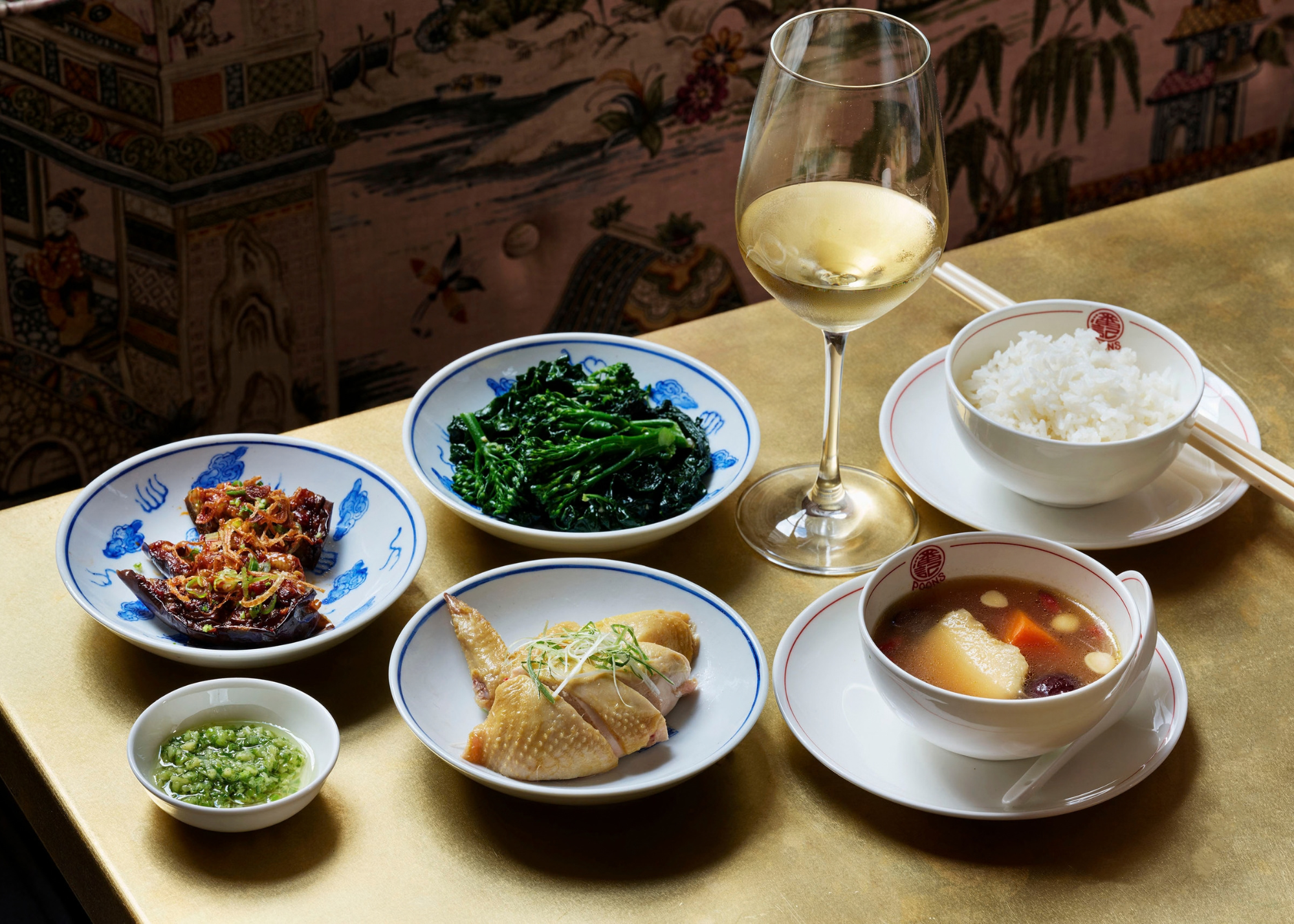 A brassy gold table with plates of chicken, soup, broccoli, and rice, beside a glass of white wine