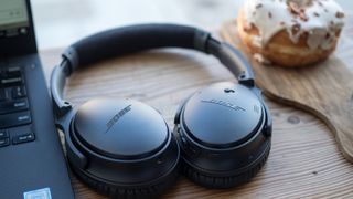 Bose QuietComfort 35 II på et træbord ved siden af en laptop og en donut