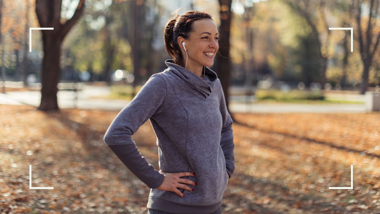 Woman standing smiling in a leafy forest with hands on hips listening to music through headphones, representing is walking good exercise