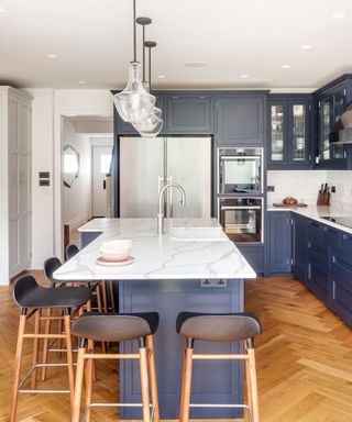 A kitchen with a white marble kitchen island with an indigo blue base, five black stools with wooden legs around it, three glass pendant lights hanging from the ceiling, and blue cabinets to the right