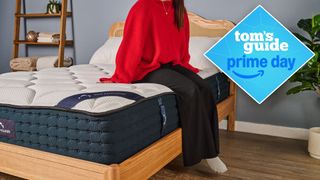 A close up of a person sitting on the edge of the DreamCloud Hybrid Mattress on a bed frame in a bedroom, a Tom's Guide Prime Day deals graphic in the top right corner
