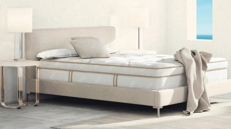 Memory foam vs latex mattresses: Image shows the Saatva Latex Hybrid Mattress on a light coloured bedframe, and covered with a natural colour bed throw