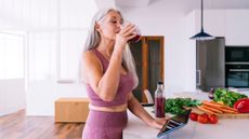 Woman in sportswear stands and drinks beetroot juice from a glass in front of a kitchen counter