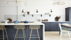 A navy blue kitchen with an island in the middle and bar stools beside the island. Above the island is a large pendant light. You can see a view of the open shelving on one side of the kitchen too as well as the corner of the dining table. 