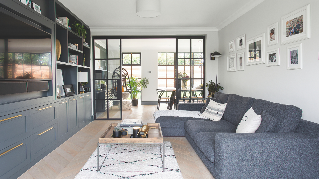 Dark blue sofa and fitted cupboards, pale rug, cushions and fabric throw, view through to the dining area with hanging chair and dining table, French doors to the patio. Gemma and Navin Mahabeer&#039;s 1930&#039;s five bedroom family home in Buckinghamshire, renovated and restyled