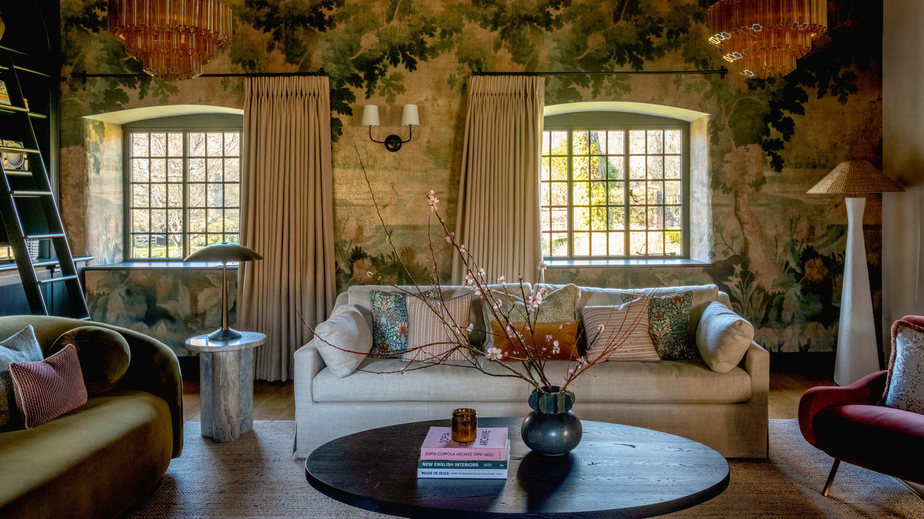 large living room with painterly style botanical mural wallpaper, two large windows, built ins to the left, a neutral couch and a velvet curved couch with a black wooden rounded coffee table