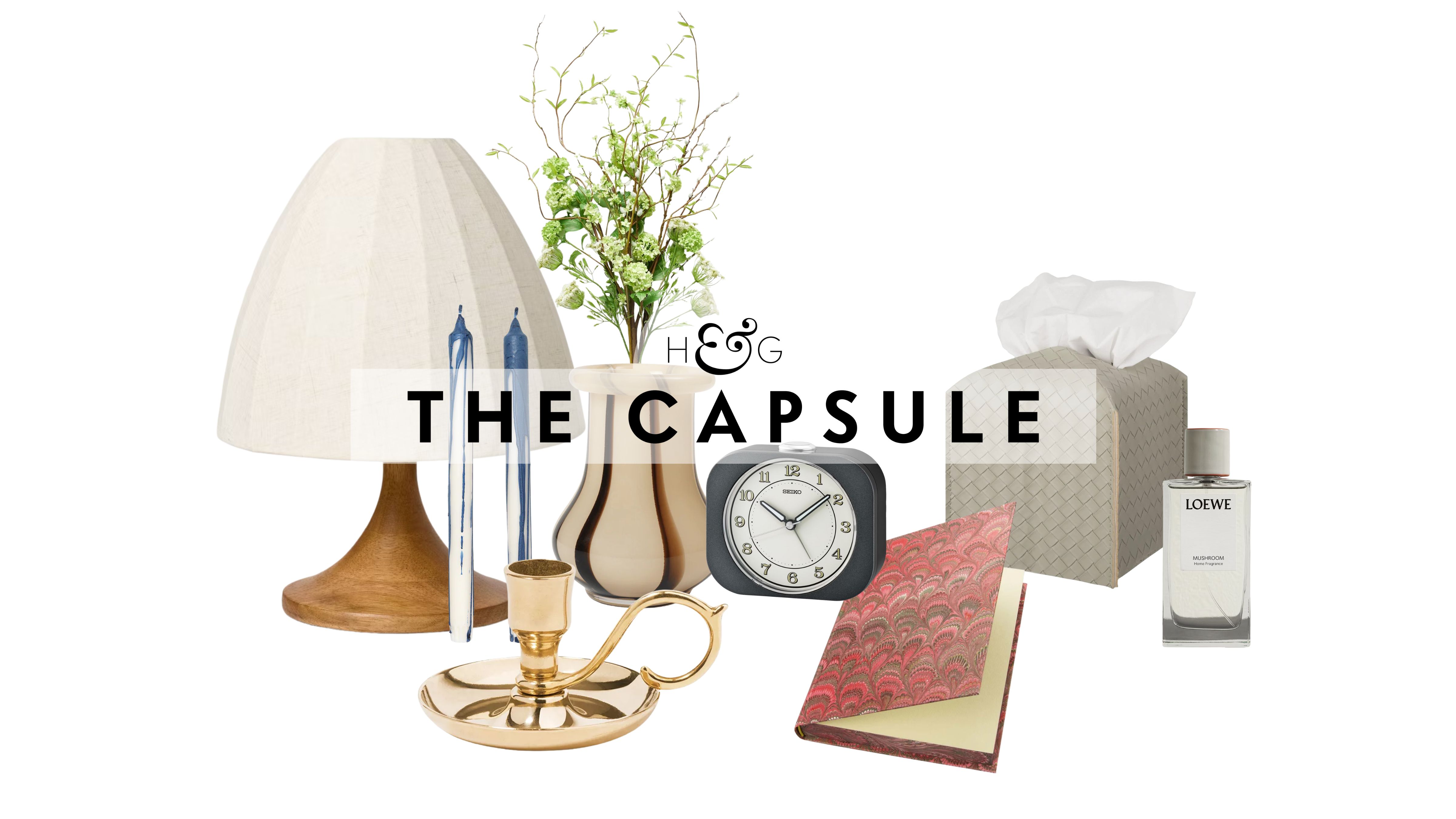 Bedside table decor pictured against a white background, including a table lamp, taper candle duo, brass candlestick, striped vase, retro-style alarm clock, patterned notebook, woven leather tissue cover, and Loewe Home spray