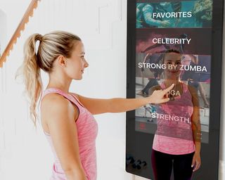 A female touching the Echelon REFLECT Touchscreen Fitness Mirror which has been installed under the stairs in the home