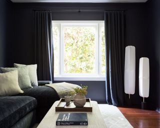 Cozy living room with black painted walls, black curtains, black sofa, decorated with cream cushions and throw, dark wooden flooring, gray rug, cream upholstered ottoman, two white lantern floor lamps
