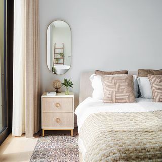 Beige bedside tables and an oval mirror hang on the wall above the bed, with decorative cushions.