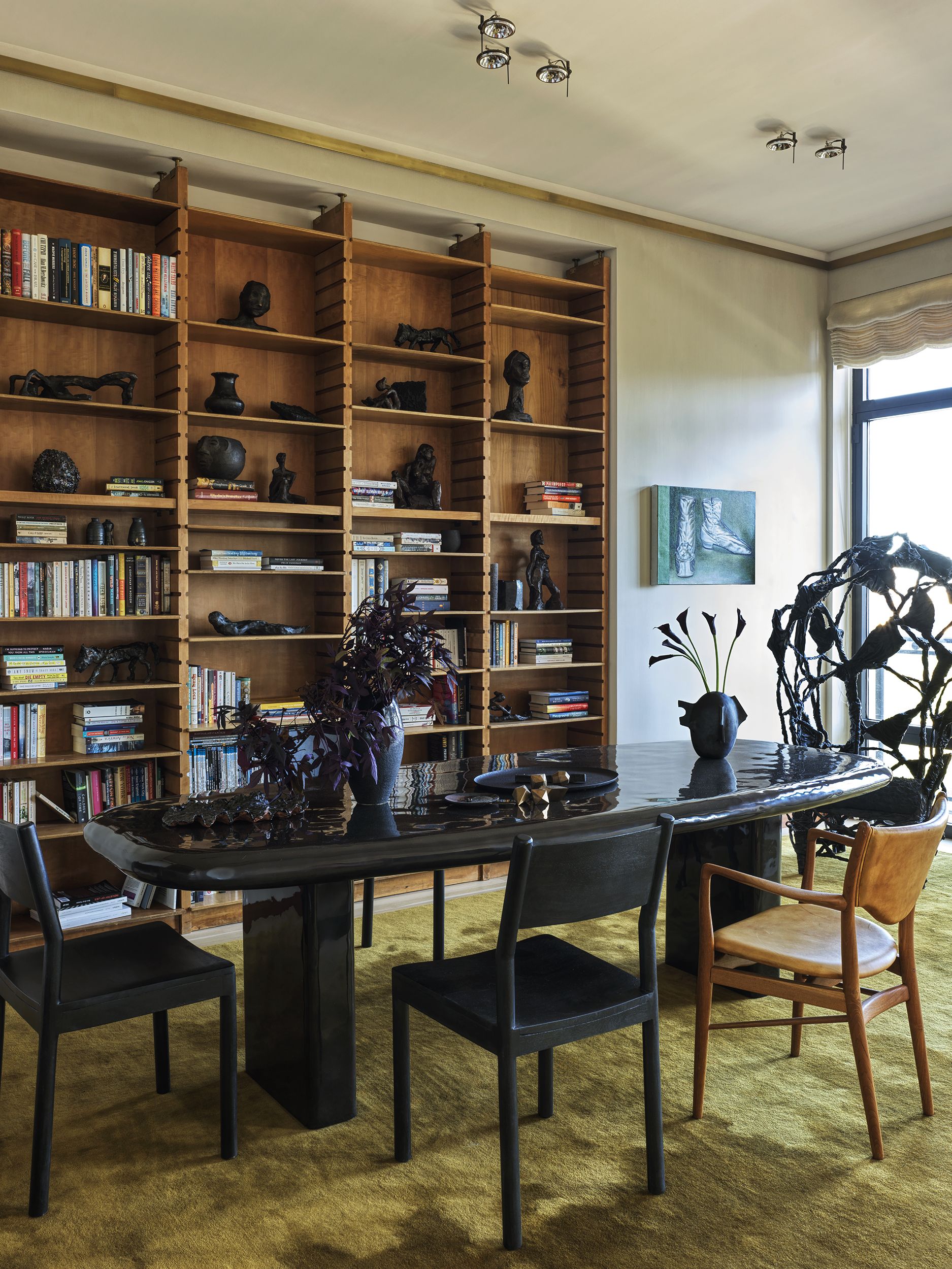 A large wooden shelving unit filled with books and decorative objects in front of a black oval dining table with black and wood dining chairs with a black sculpture in the corner of the room