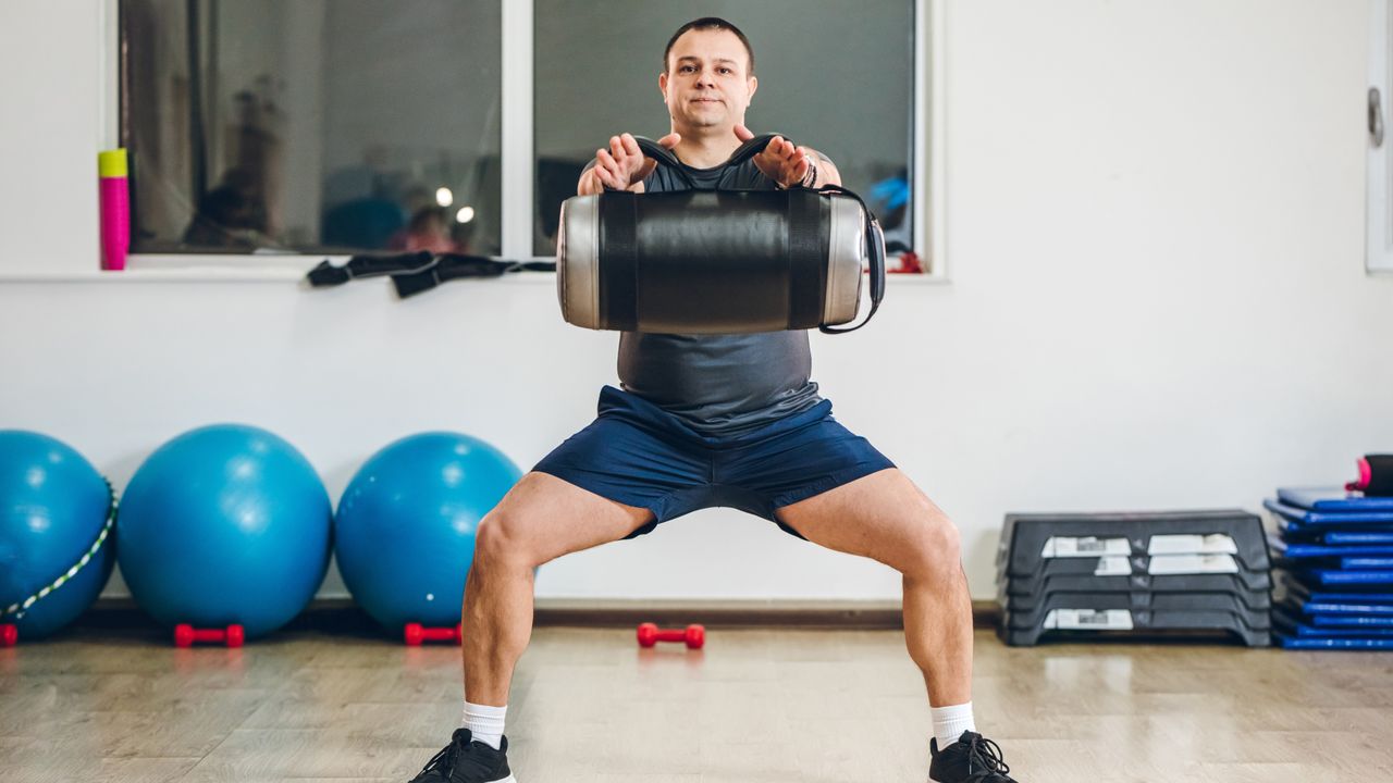 A man performs wide legged squats while holding a sandbag by its handles. His arms are straight out in front and his knees are bent and pointing outwards. Behind we see several exercise balls, gym steps and dumbbells.