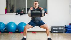 A man performs wide legged squats while holding a sandbag by its handles. His arms are straight out in front and his knees are bent and pointing outwards. Behind we see several exercise balls, gym steps and dumbbells.