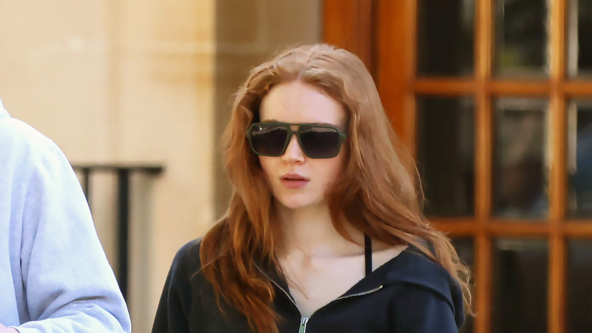 Sadie Sink wearing a black zip-up hoodie, low-rise long denim shorts, and black leather flats