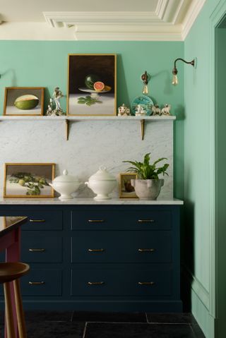 A kitchen with mint green wall paint decor, white shelf with framed wall art, traditional wall lights, marble backsplash and navy blue kitchen cabinetry