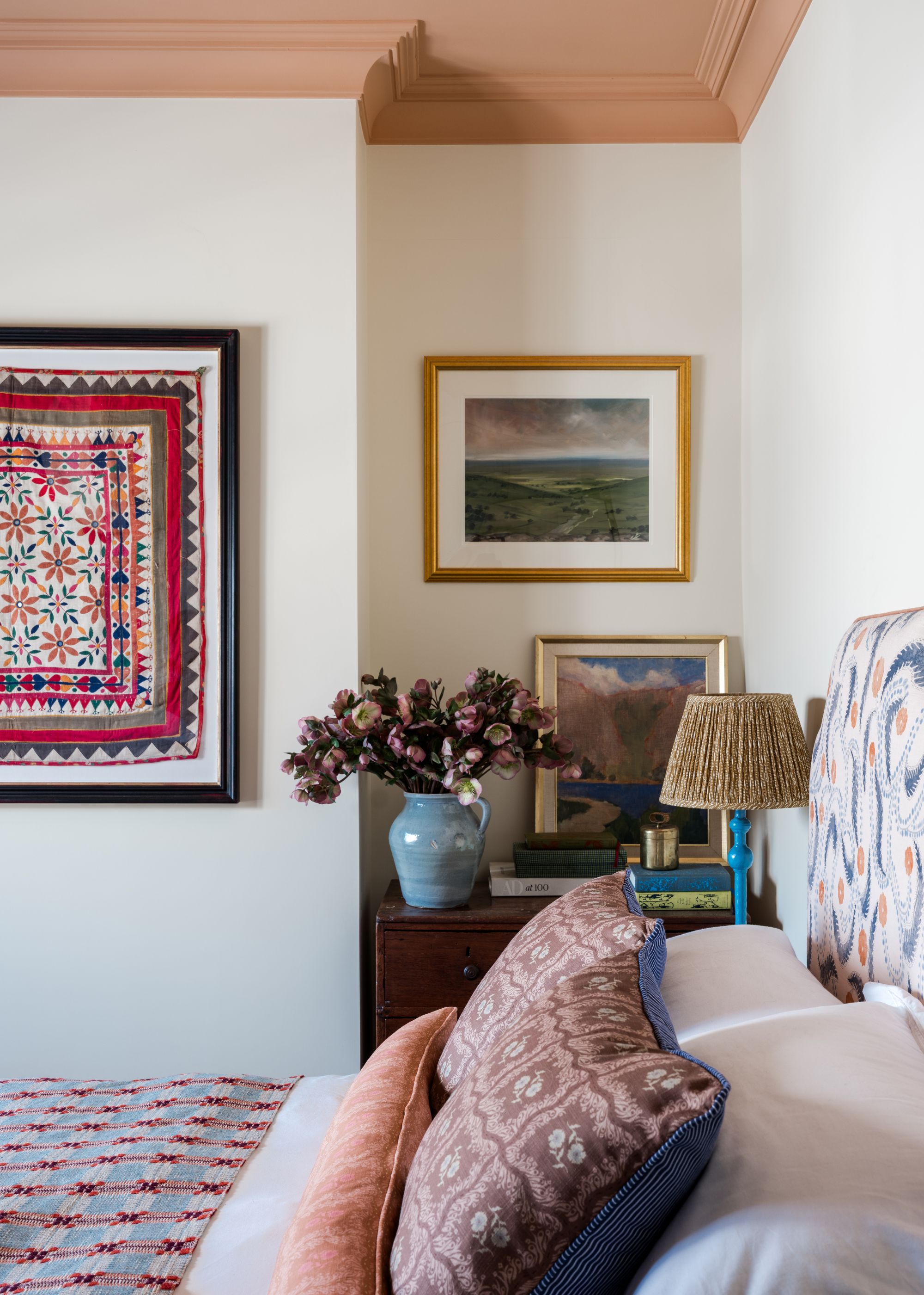 A cozy bedroom with warm white walls and a pink ceiling, a bed with muted red patterned textiles, a framed textile on the wall, along with landscape artworks, and a vase of flowers on a dresser.