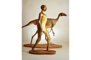 A model of a humanoid, dinosaur-like creature, with a model of a dinosaur behind it