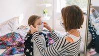 Picture of a young girl sat on a bed while her mother puts a blue surgical mask on her face. The mother is also wearing the same kind of mask. They are in a bedroom with a large window in the background, a mirror behind the mother on the right-hand side of the image and in the background there are some shelves, one of which has a pot plant on it 