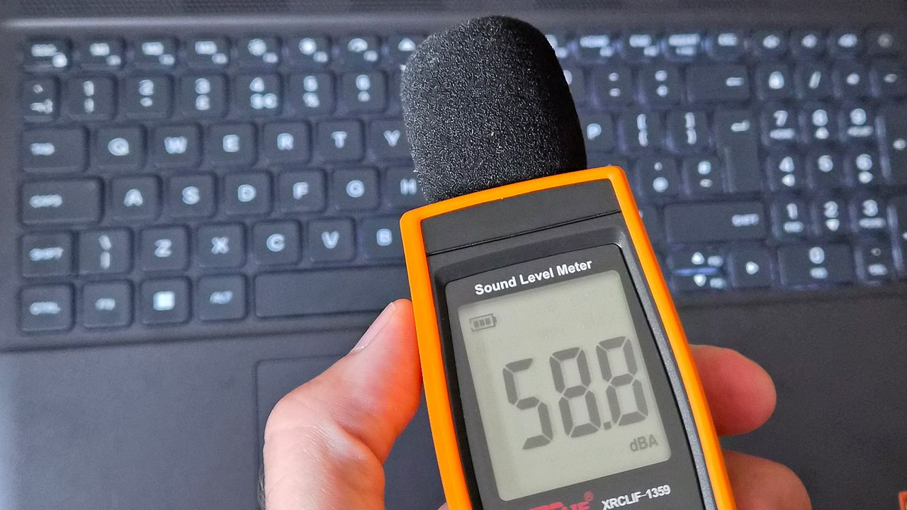 A handheld orange sound level meter with a foam‑covered microphone displays a reading of 58.8 dBA while being held above an Alienware 15 laptop keyboard.