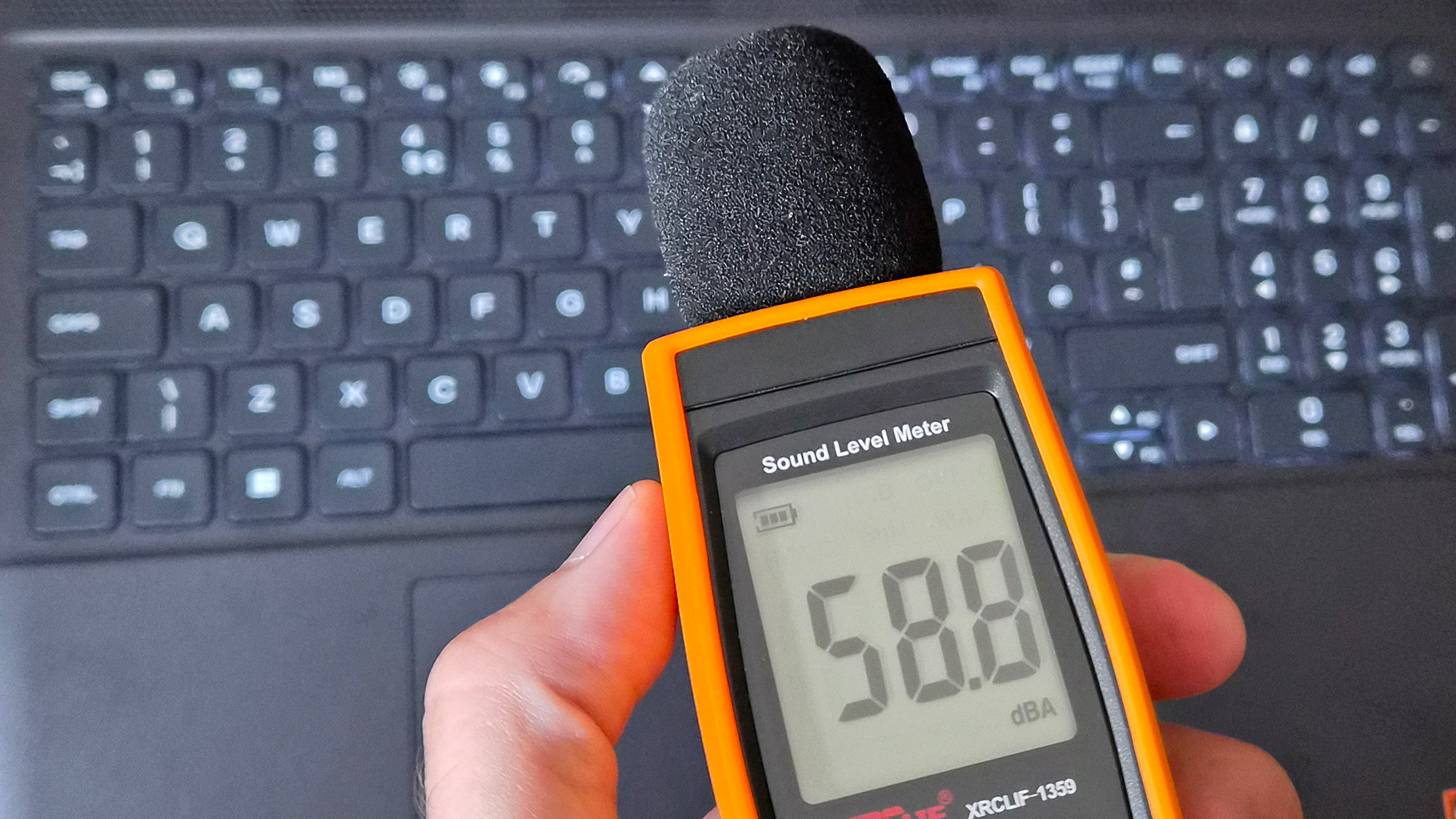 A handheld orange sound level meter with a foam‑covered microphone displays a reading of 58.8 dBA while being held above an Alienware 15 laptop keyboard.