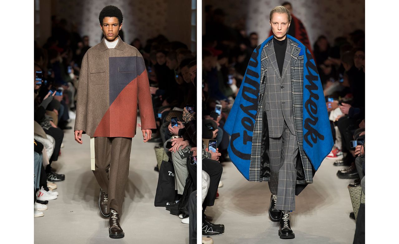 Catwalk view of two models wearing looks from OAMC&#039;s collection. One model is wearing a white top, brown, blue and red jacket, brown trousers, white belt and brown boots. And the second model is wearing a black top, grey grid style suit, grey plaid coat, blue shawl with black writing and black boots. There are people seated on both sides of the catwalk