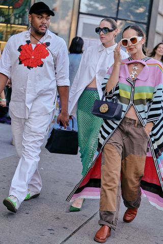 Designer Christopher John Rogers in a white outfit walking the street at New York Fashion week two women in a white button down, green skirt, colorful striped sweater, brown pants, and a cookie bag