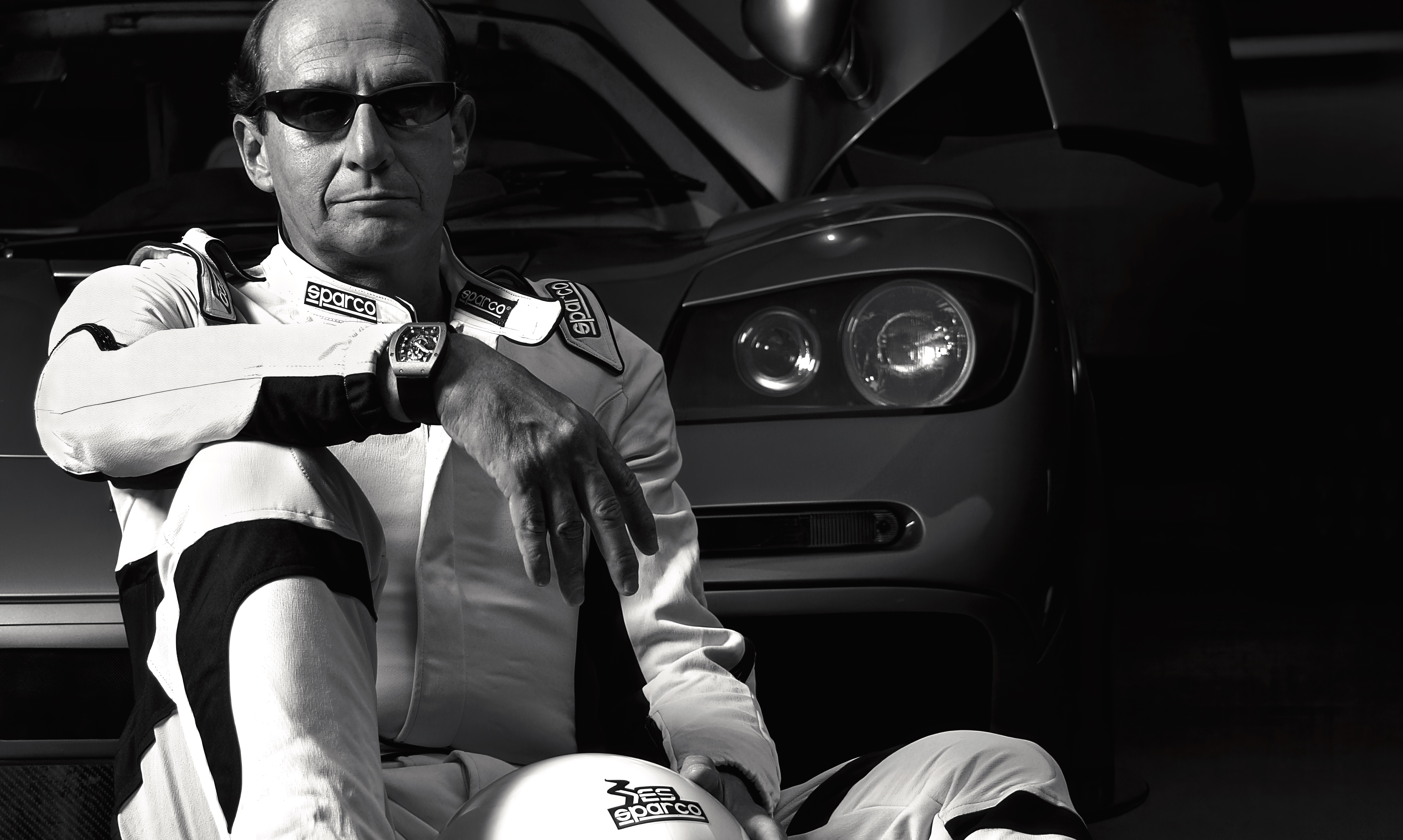 Black and white image of a man in racing overalls sitting in front of a car