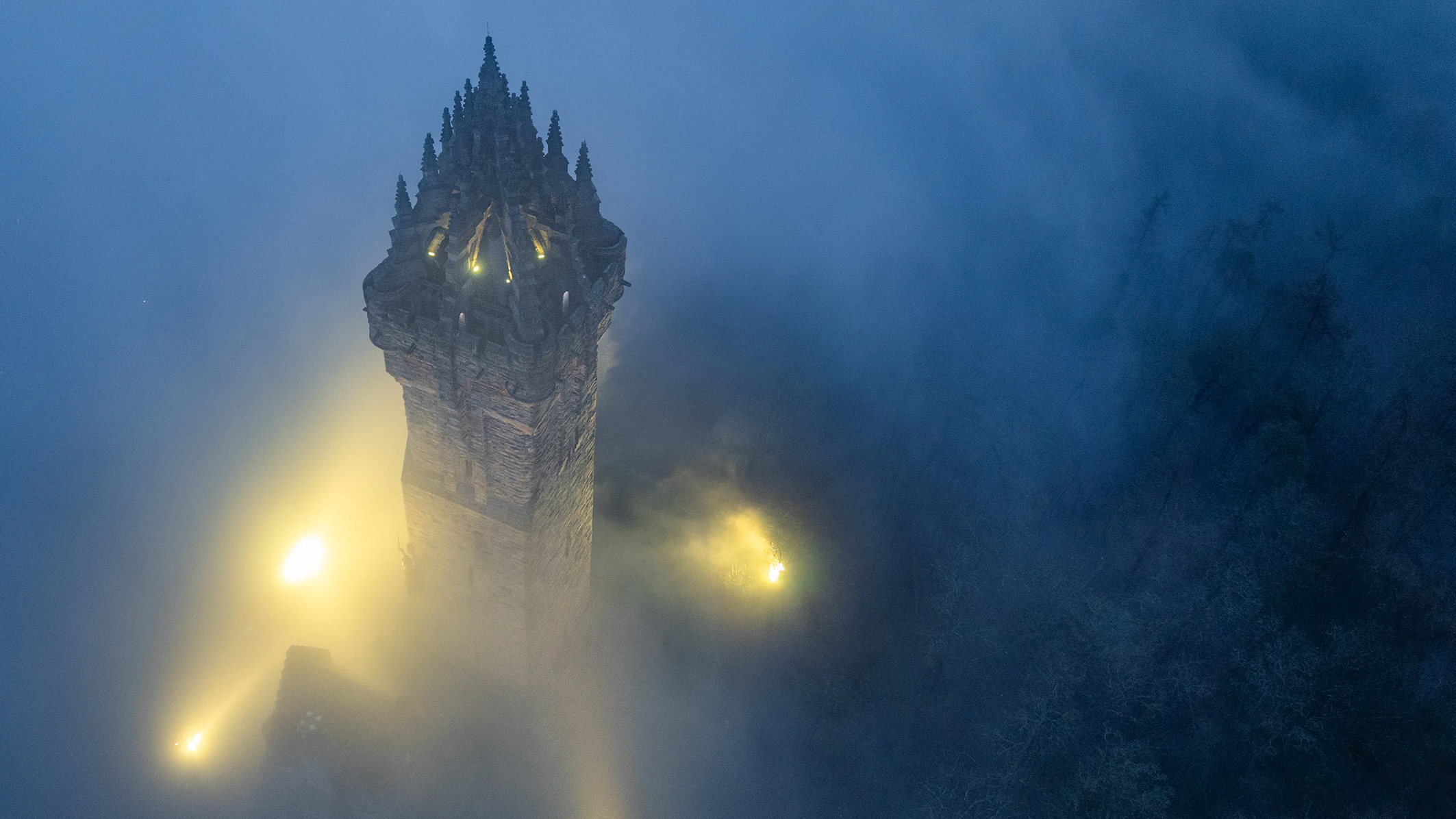 Aerial view of a tall stone tower emerging from thick fog, illuminated by soft yellow lights, surrounded by shadowy trees