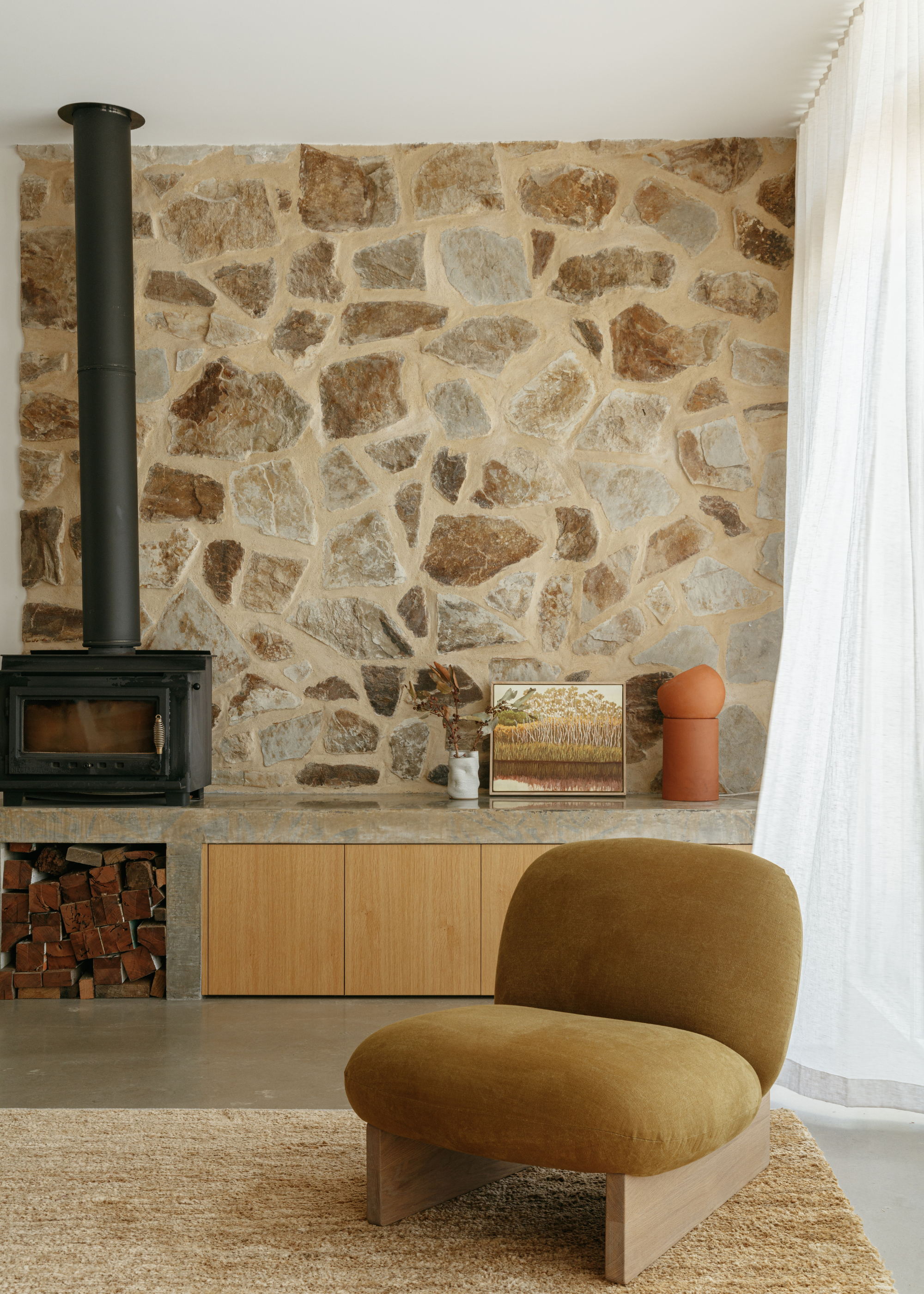 A calm minimalist living room with a mustard sofa seat, a wheat-colored area rug, white window treatments, a stone wall, a black fireplace, a stack of wood logs, an orange lamp