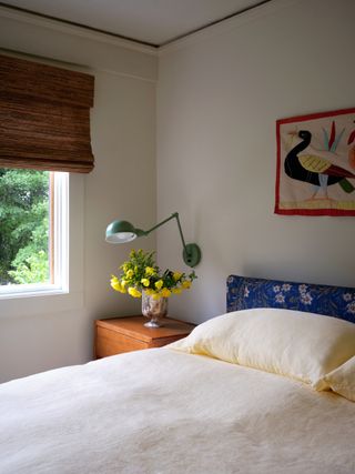 small bedroom with cream sheets, upholstered bedhead, side table with flowers, and bamboo blinds with pelmet