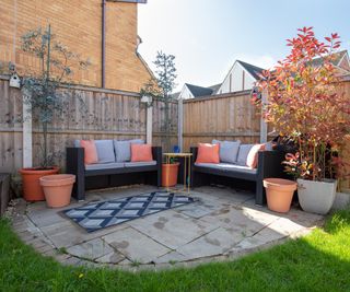 small round patio area in corner of garden with grey slabs, rattan furniture and plant pots with small trees