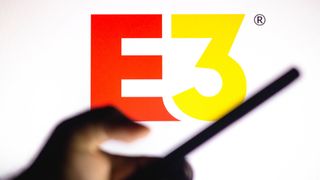 En suddig hand håller upp en mobil framför en stor E3-logo i rött och gult som visas upp mot en vit bakgrund.