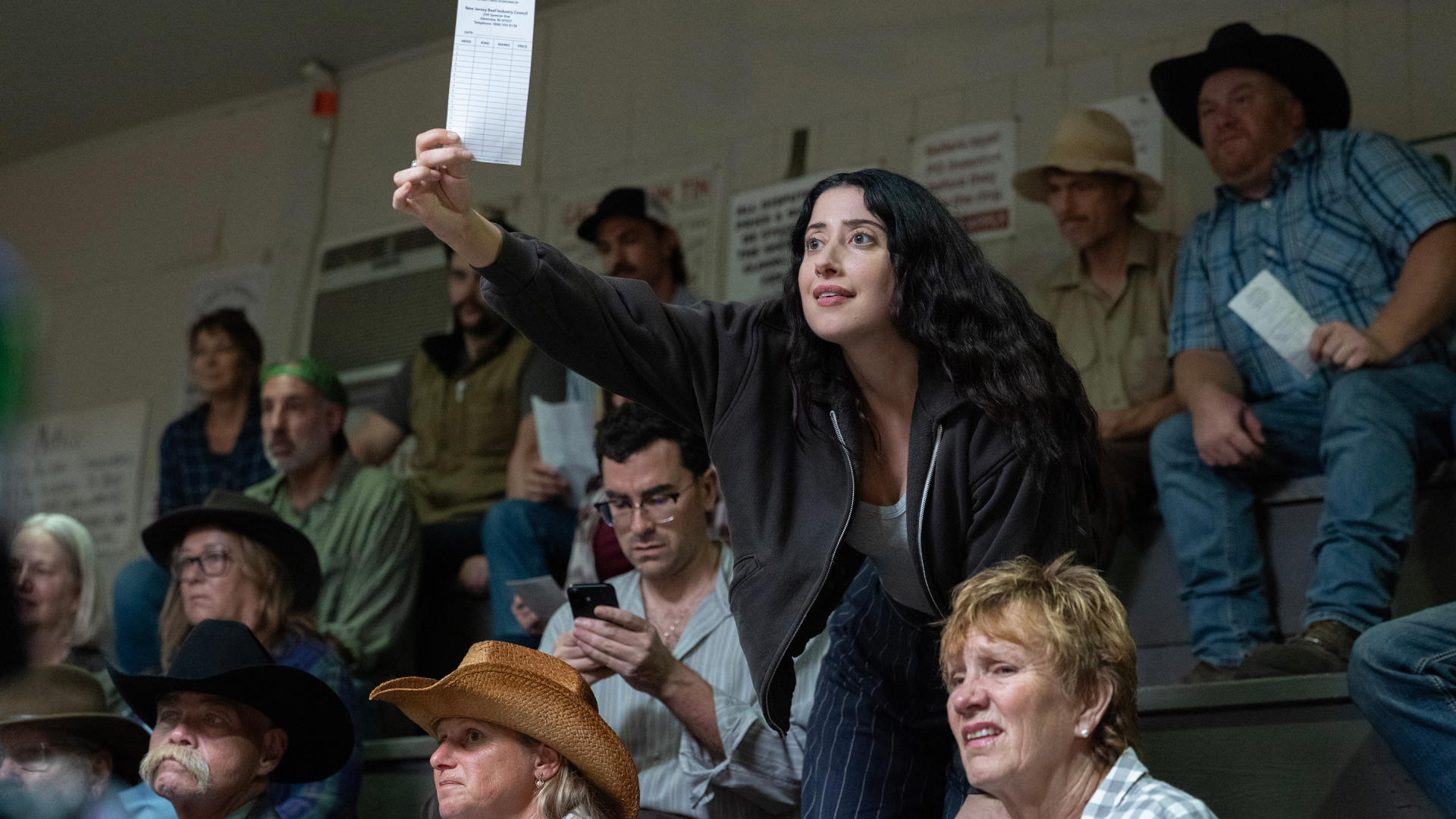 A man (Dan Levy as Nicky) looks down at his phone and a woman (Taylor Ortega as Morgan) stands while holding up a bidding slip, as they sit in a crowd at an auction, in Episode 105 of BIG MISTAKES.