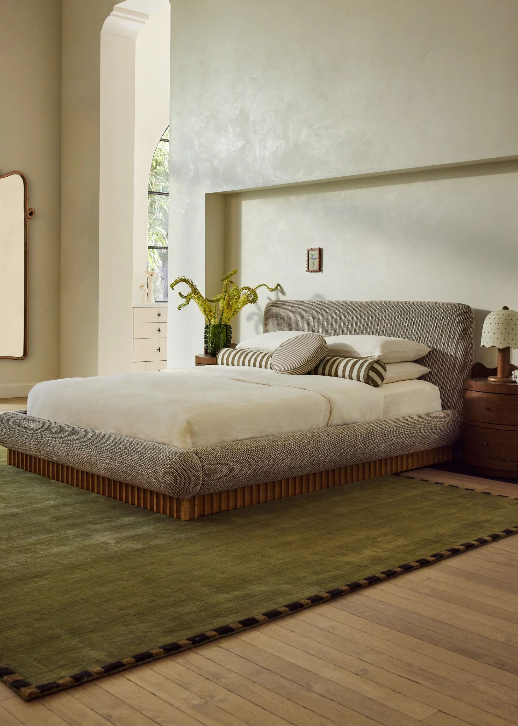 A minimalist bedroom with off-white walls, a gray bed with a wood base and a white bedscape, a vase with fuzzy stems, and a green area rug