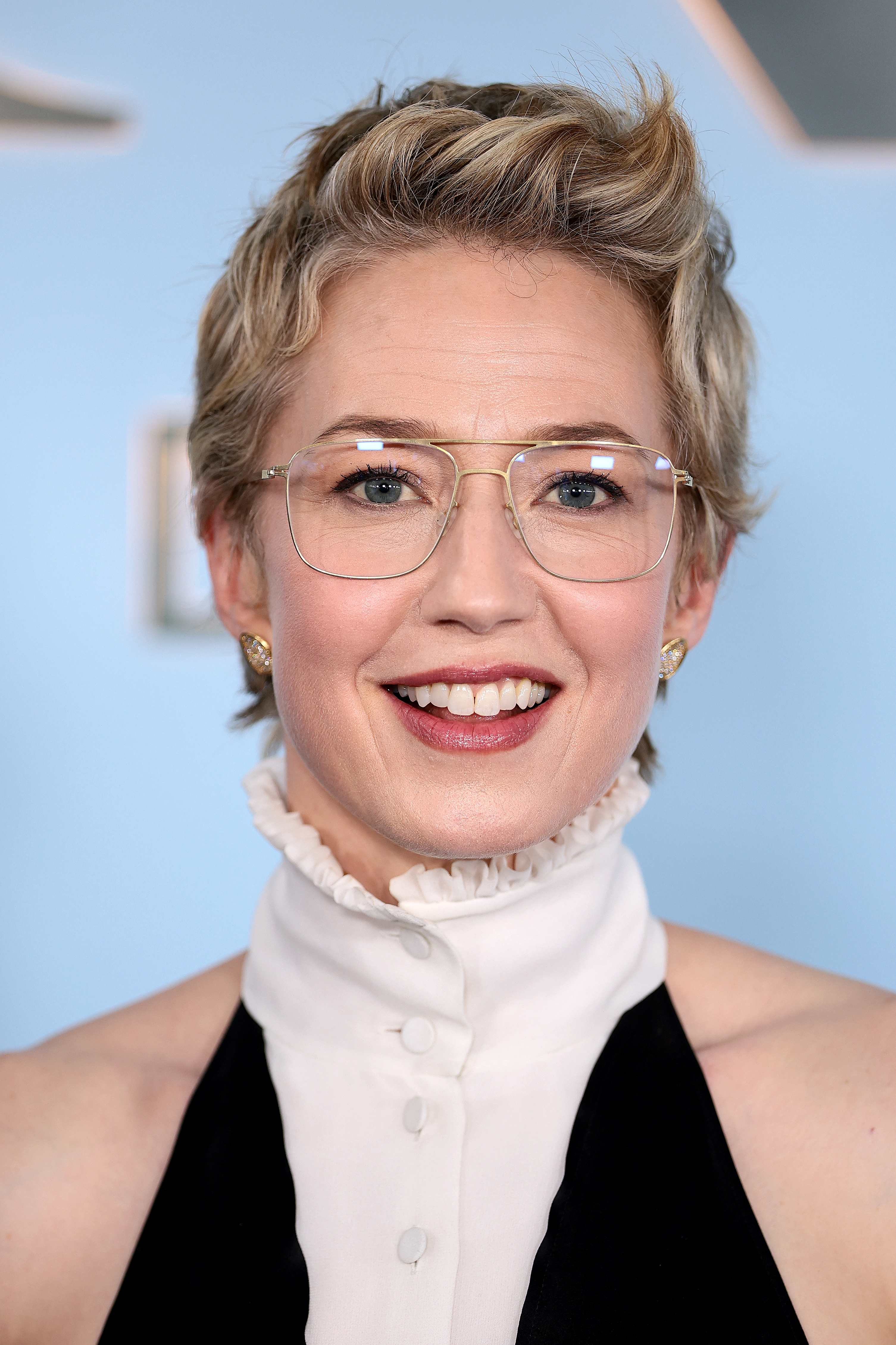 a white woman with glasses and short, blonde hair wears a white turtleneck and poses on a red carpet