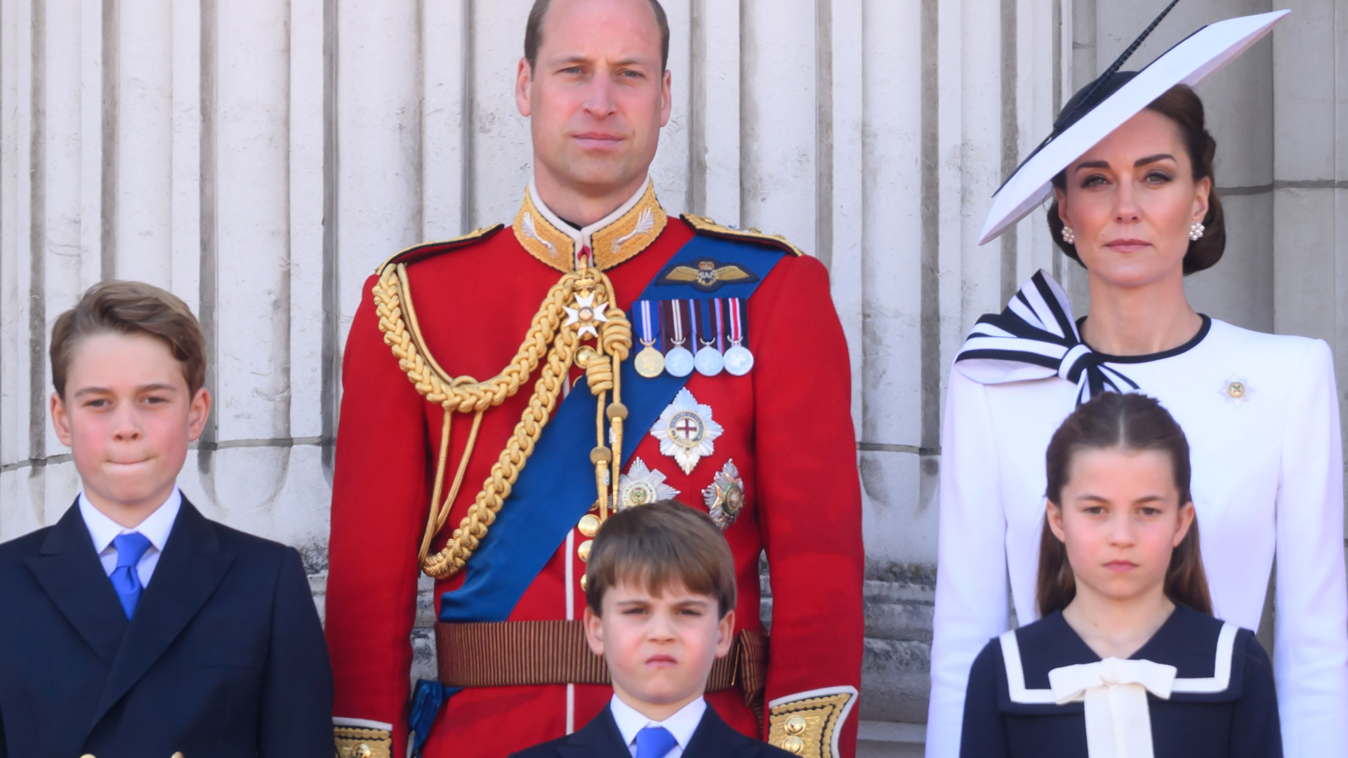 Prince William wears red military uniform and Kate Middleton wears a white dress with navy detailing, as they stand on the Buckingham Palace with their three kids, Prince George, Prince Louis, and Princess Charlotte